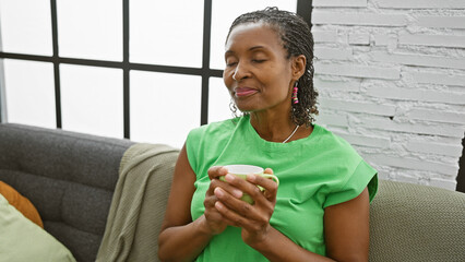 A content african american mature woman enjoys a cup of coffee in a cozy living room setting.
