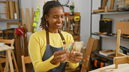 African american woman smiles holding money in a carpentry workshop