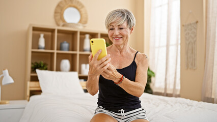 A mature woman interacts with a smartphone in her cozy, well-decorated bedroom, showcasing technology usage among adults.