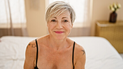 A mature woman with grey short hair smiles warmly in a bedroom setting, exuding elegance and tranquility.