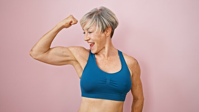 A fit, mature woman with short grey hair flexes her muscles against a pink isolated background, displaying strength and vitality.