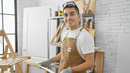 A young hispanic man smiles in a well-lit carpentry workshop wearing safety glasses and an apron