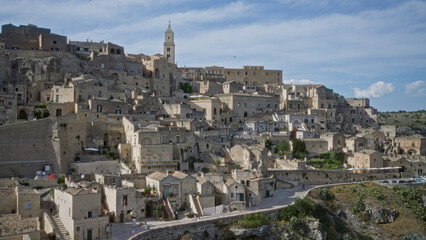 Obraz premium Historic townscape of matera in basilicata, italy with ancient stone buildings, narrow streets, and a prominent bell tower under a blue sky.