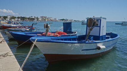 Fototapeta premium Fishing boats docked at the harbor in porto cesareo, italy, with a clear blue sky and coastal town in the background.