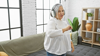 Senior woman with grey hair dancing in a living room, wearing headphones and casual clothes,...