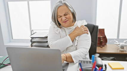 Mature grey-haired woman grimacing in pain at office due to shoulder ache
