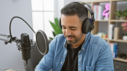 Hispanic man smiles while recording in a modern radio studio with professional microphone and...