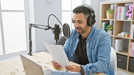 Handsome hispanic man recording audio in a modern radio studio with microphone and laptop