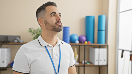 A thoughtful hispanic man with a beard stands in a healthcare clinic interior, conveying a...