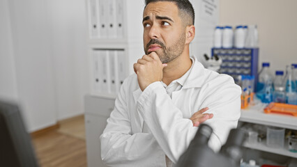 Pensive hispanic man in lab coat contemplating in a laboratory filled with chemical bottles.