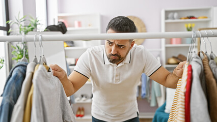 Hispanic man perplexed by clothing choices in a bright, indoor wardrobe room.