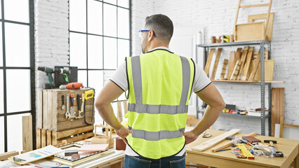 Back view of a young hispanic man with safety vest and glasses in a bright carpentry workshop.