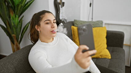 Young hispanic woman vaping indoors, casually using her phone in a cozy living room setting.