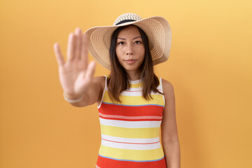 Middle age chinese woman wearing summer hat over yellow background doing stop sing with palm of the hand. warning expression with negative and serious gesture on the face.