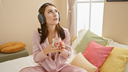 A contemplative young woman in pink pajamas enjoys music with headphones while holding a coffee mug...