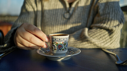 Close-up of a woman's hand holding a decorative turkish coffee cup outdoors, with soft lighting.