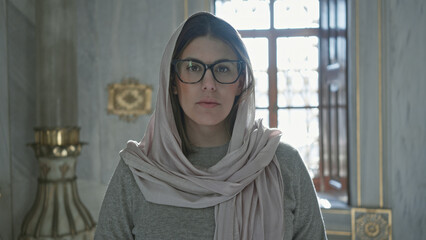 A thoughtful woman wearing glasses and a hijab poses inside a mosque, embodying a serene religious atmosphere.