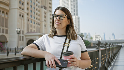 Young woman with glasses holding coffee, phone on a sunny doha skyline bridge