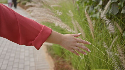A young adult hispanic woman walks through a park, brushing her hand against tall grass.