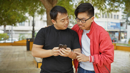 Two asian men are closely interacting with a smartphone on a sunny park bench, exemplifying...