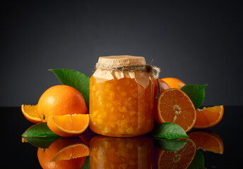 Orange jam in glass jar and fresh fruits on a black reflective background.