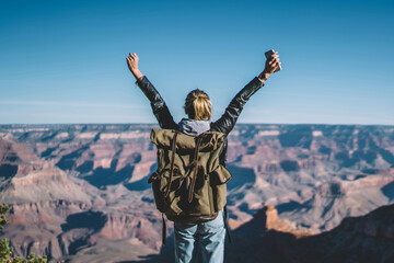 Fototapeta premium Back view of young wanderlust feeling victory conquering mountain in Grand Canyon, female traveler with backpack raising hands enjoying beautiful nature from high rocky hills getting to destination