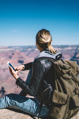 Rear view of female traveler checking connection on telephone sitting on high mountain hill, hipster girl wanderlust satisfied with data network in wild environment holding smartphone for chatting