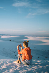 Young woman using smartphone for making photo of desert dunes during sunset having journey to White sands park, hipster girl taking pictures of scenic nature of scenery lands in famous landmark in USA