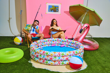 Couple creates beach party sitting, sunbathing and relaxing by inflatable kiddie pool and toasting with drinks. Concept of pop art, remote work, travelling, recreation, lifestyle, fashion and style.