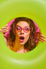Portrait of surprised young woman holds neon-green inflatable ring, peeking out of it against bright pink background. Concept of pop art, remote work, party, recreation, lifestyle, fashion and style.