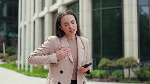 Unhealthy pretty caucasian female feeling chest pain while standing in middle of street with modern smartphone in hands. Sorrowful young woman touching body and trying to easing ache outdoors.