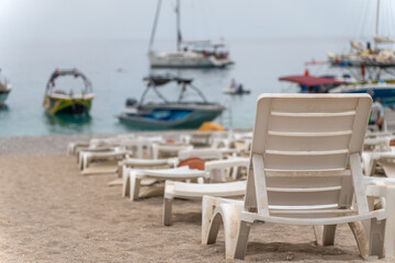 Close-up of white plastic sunbeds on the beach against the background of the blurred sea, mountains, yachts and boats.