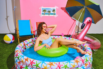 Beautiful woman relaxes in inflatable pool with green float ring against pink backdrop. Fun and...