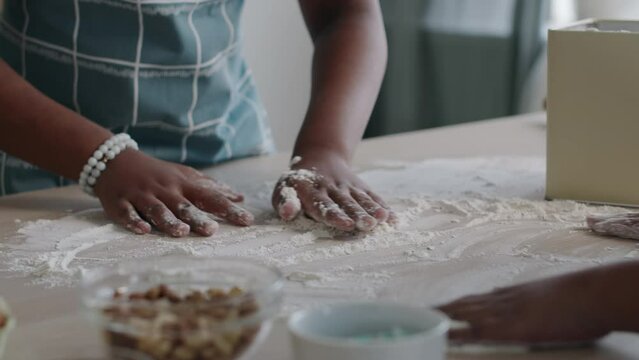Close-up of hands of unrecognizable black woman and children dusting flour over kitchen table surface going to prepare dough-based dish or knead pastry for bread