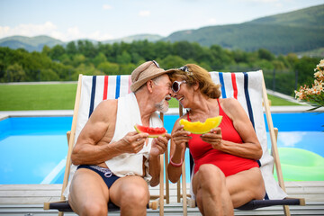 Beautiful elderly couple enjoying their vacation. Seniors having romantic time, sitting by swimming pool in lounche chairs sunbathing.