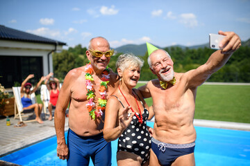Group of cheerful seniors standing by pool taking selfie. Elderly friends spending hot day by swimming pool, sunbathing.
