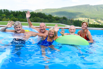 Group of cheerful seniors shaving fun in pool jumping, swiming and lounging on floats. Elderly friends spending hot day by swimming pool.