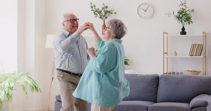 Elderly man leads his wife in dance, having fun together in bright living room of his cozy home. Cheerful Caucasian mature family energetically spending time together. Concept of active elderly people