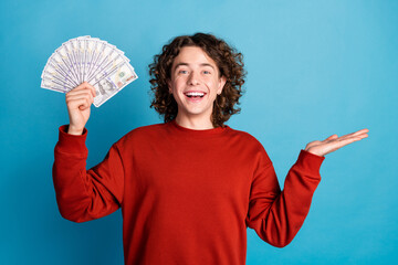 Photo of handsome excited guy wear red sweater showing dollars fan showing arm emtpy space isolated...