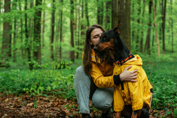 young woman walking with her doberman dog in the forest. Wearing yellow rain jackets girl and dog playing outdoors. Human and dog friendship concept