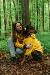 young woman walking with her doberman dog in the forest. Wearing yellow rain jackets girl and dog playing outdoors. Human and dog friendship concept