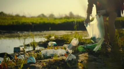 Cleaning the shore of a pond, young man carefully collects plastic waste. It sorts waste plastic bottles and puts them into garbage bag for further recycling, maintaining sustainable recycling process