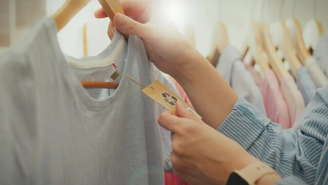 Clothes recycling. The look of a modern girl, her gaze fixated on hangers with clothing reuse signs in a stylish accessories store. A woman following fashion trends holds a tag with a recycling symbol