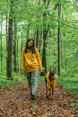 young woman walking with her doberman dog in the forest. Wearing yellow rain jackets girl and dog playing outdoors. Human and dog friendship concept