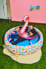 Young businessman in formal suit relaxing in kiddies swimming pool on flamingo inflatable-ring and drinks beer. Concept of pop art, party, recreation, lifestyle, fashion and style.