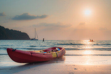 Fototapeta premium Tranquil beach scene at sunset with a red kayak on the shore, a sailboat, and kayakers in the distance.