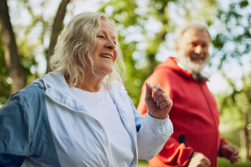 Focus on happy smiling elderly woman in sportswear running with man in public park on warm early...