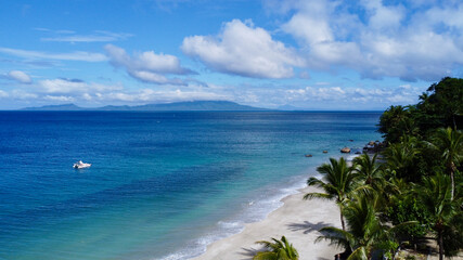 Aerial view of the coast of a tropical island. Top view of a sandy beach with palm trees, the surface of a calm sea, clouds in the sky and a small boat in the sea.