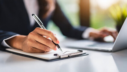 
A businesswoman's hand holding a pen, poised to write on a form atop a white table. The macro focus highlights the meticulousness and attention to detail required in business documentation