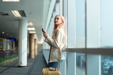 Young woman with suitcase using smartphone at airport terminal. Modern travel concept, casual outfit, waiting for flight